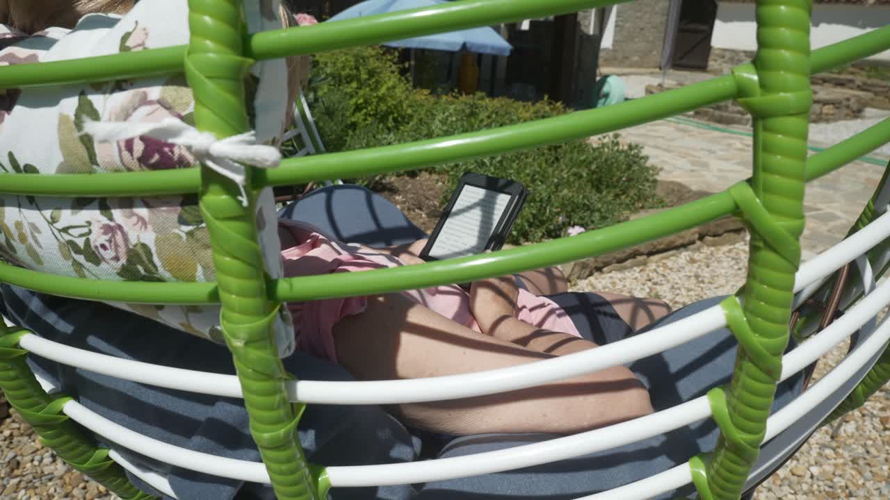 Woman sits in comfortable garden swing chair reading eBook novel on warm sunny day. slow push shot over shoulder view