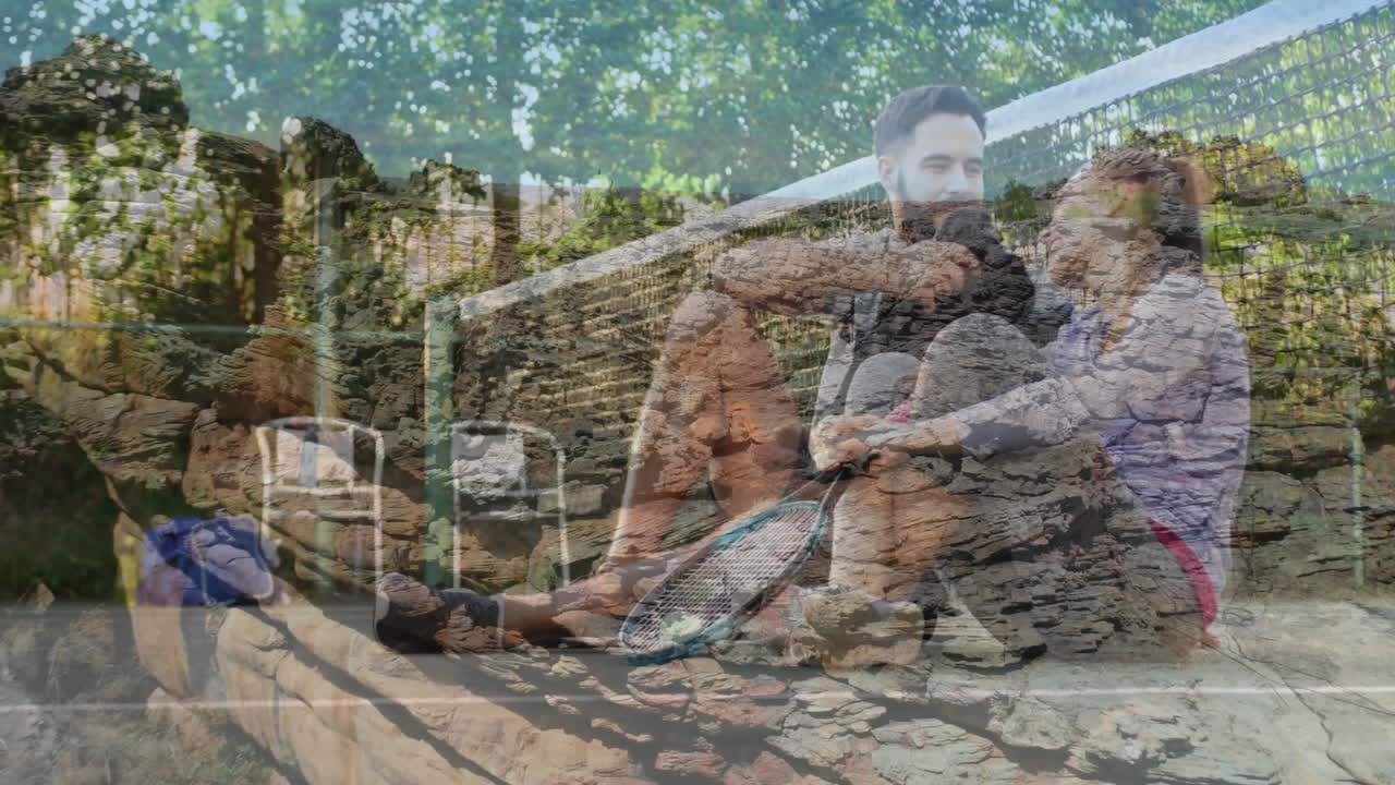 Holding tennis rackets, couple sitting against stone wall with trees in background