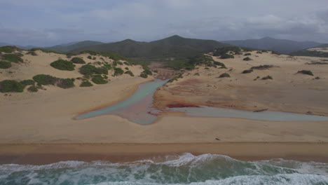 Spiaggia-di-Piscinas,-Sardinia:-flying-from-the-sea-towards-the-shore-of-the-beach-and-seeing-the-canals-that-connect-sea-and-land