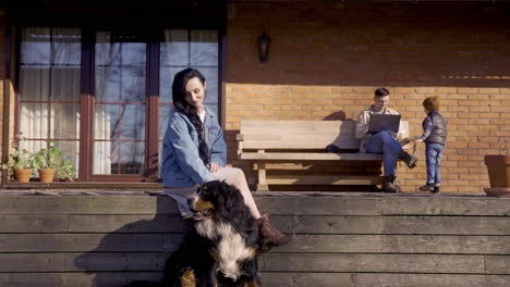 Caucasian-woman-petting-her-dog-while-her-partner-is-using-a-laptop-and-playing-with-his-son-outside-a-country-house