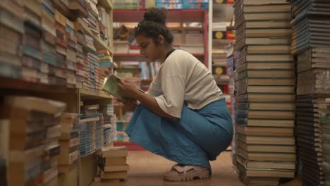 Asian-girl-sitting-and-holding-a-book-with-thoughtful-facial-expression-in-the-middle-of-bookshelves,-Side-angle-shot