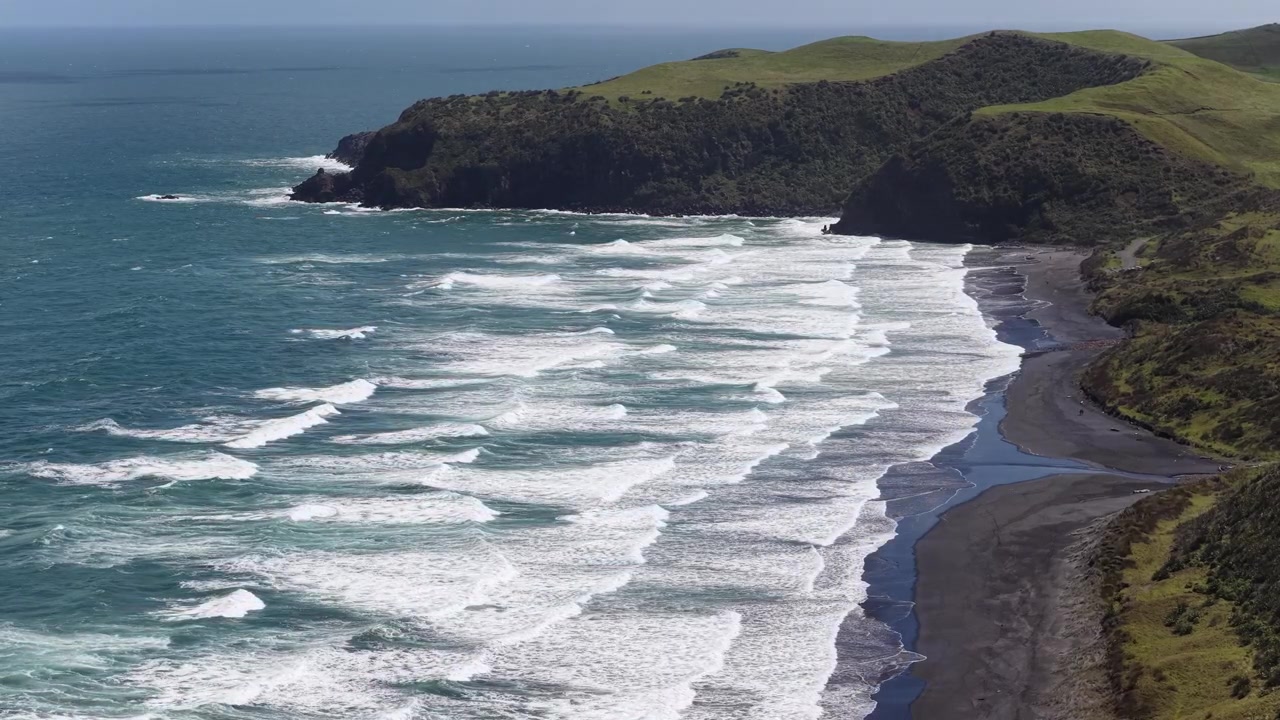 Drone flight above the wild black sand coastline of New Zealand's North Island, revealing powerful Pacific surf, rugged cliffs, rolling green hills, and an unspoiled volcanic shoreline.