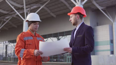 Joven-Gerente-Del-Proyecto-Con-Traje-Negro-Que-Examina-El-Objeto-De-Construcción-Con-Un-Trabajador-De-La-Construcción-Con-Uniforme-Naranja-Y-Casco