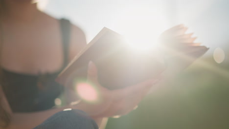 partial view of woman seated outdoors holding book as she flips through, wind blows pages creating movement, sunlight creates warm ambience around book