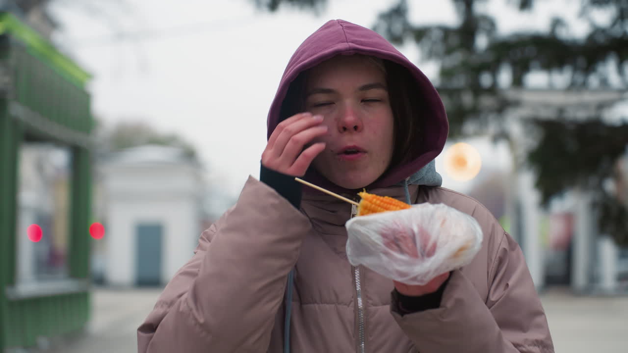 겨울 재을 입은 소녀가 거리에서 옥수수를 먹고, 거리 음식을 즐기고, 추운 날씨, 플라스틱으로 싸여, 도시 환경, 야외에서 캐주얼 식사, 옥수수, 후드 재, 음식 즐거움
