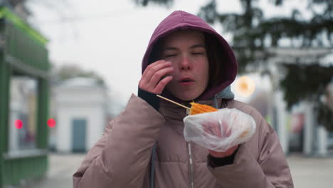 chica con chaqueta de invierno comiendo maíz en la calle, disfrutando de la comida callejera, clima frío, envuelta en plástico, entorno urbano, comida casual al aire libre, sosteniendo maíz, chaqueta con capucha, disfrute de la comida