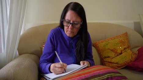 Middle-aged-Woman-Taking-Notes-During-Online-Class-On-The-Couch-Inside-Her-House