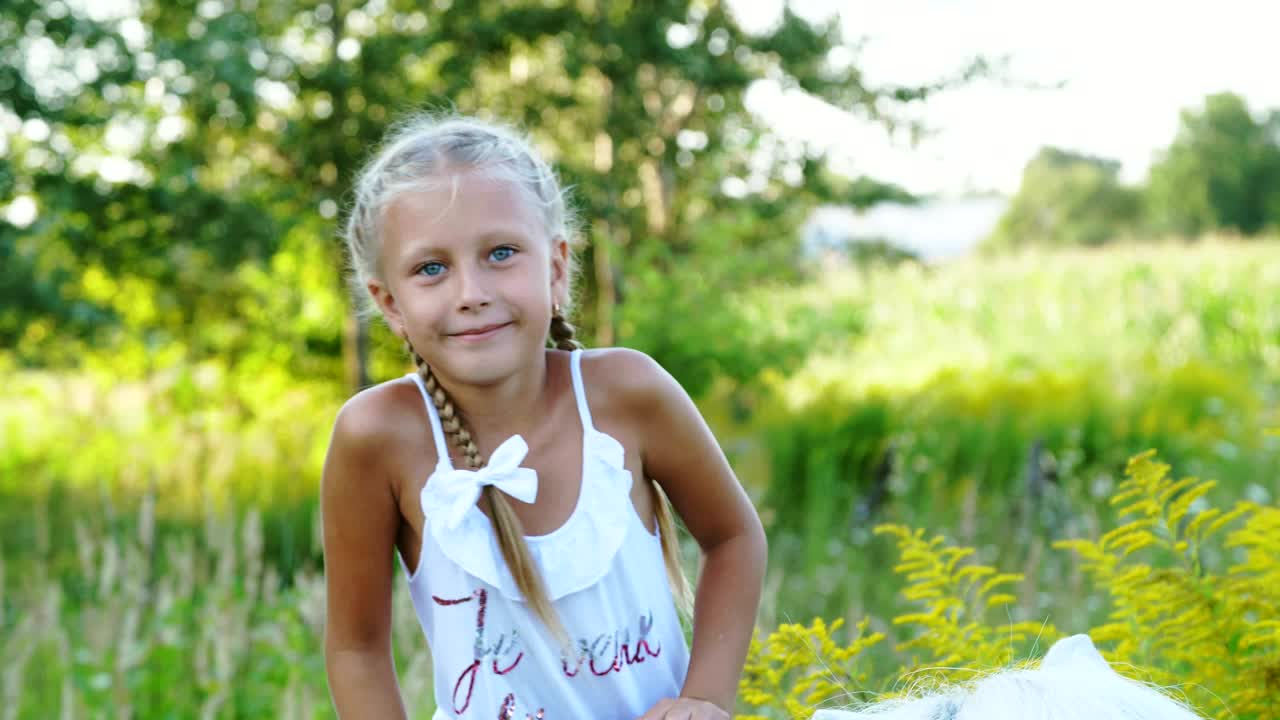 Siebenjähriges blondes Mädchen in weißem Kleid klettert auf ein Pony und sitzt darauf. Fröhlicher, glücklicher Familienurlaub. Draußen, im Sommer, nahe dem Wald.