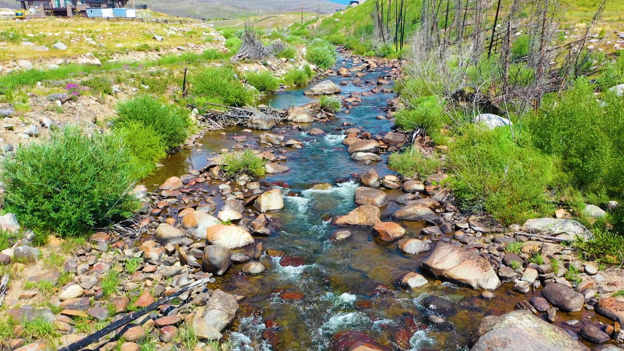 Low reverse drone flight over an Iceland style highland stream winding through stark open terrain with rocky banks low vegetation expansive valley floor and soft blue ridges beyond