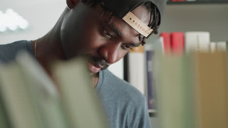 Close up of young man standing near bookshelf in library, looking focused and thoughtful while scanning books, with blurred foreground and soft background