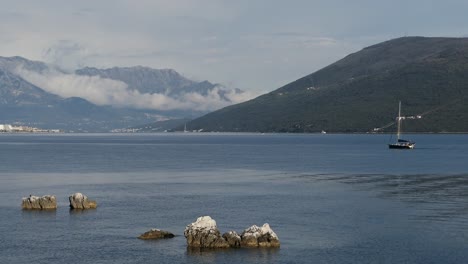Calm-sea-with-sailboat-and-clouds-around-mountains,-Kotor-Bay-area,-Montenegro