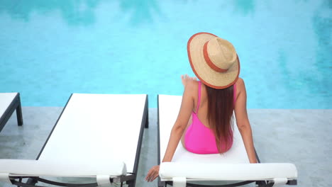 Back-view-of-young-woman-with-large-hat-and-swimsuit-sitting-on-deckchair-by-swimming-pool