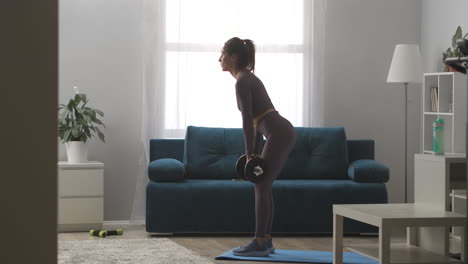 Entrenamiento-En-Casa-Con-Pesas-Joven-Mujer-Sexy-Está-Sosteniendo-Pesas-En-Las-Manos-E-Inclinándose-Hacia-Adelante-En-La-Sala-De-Estar-Estilo-De-Vida-Saludable-Y-Entrenamiento-De-Poder-De-Bienestar