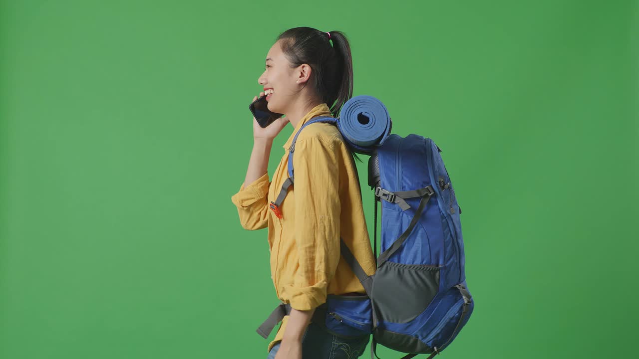 Side View Of Asian Female Hiker With Mountaineering Backpack Talking On Smartphone And Looking Around While Walking On Green Screen Background In The Studio