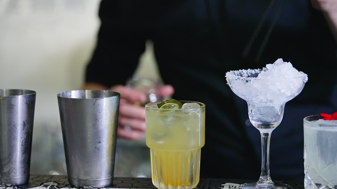 Making cocktails on the bar stand. Male bartender putting a green leaf into a glass filled with ice and beverage. Close up. Blurred backdrop.