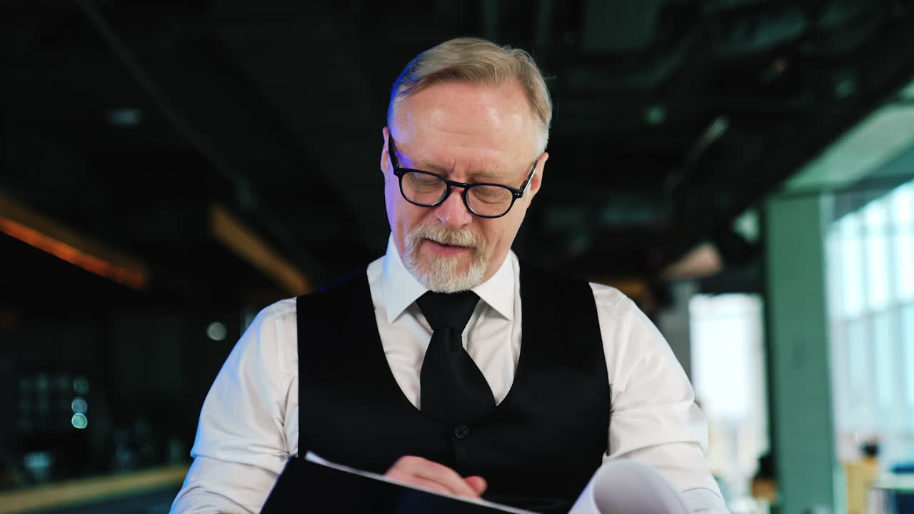 Senior fair-haired bearded man wearing glasses, shirt, tie and vest. Businessman turning pages checking information.