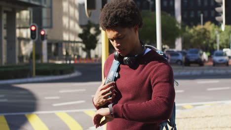 african american man in city checking smartwatch, wearing headphones and backpack waiting in street