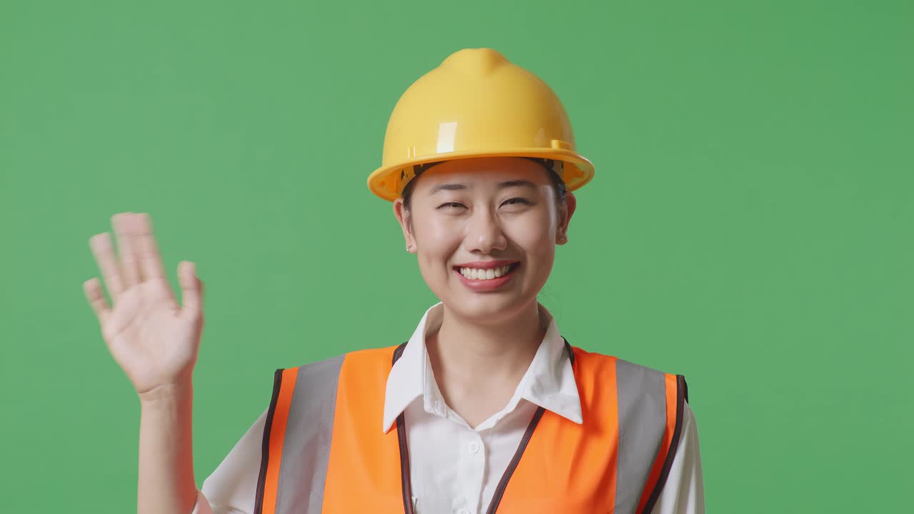 안전 헬을 입은 아시아 여성 엔지니어가 카메라에 미소 짓고 손을 흔들며 녹색 스크린 배경 스튜디오에 서서 작별 인사를 하고 있습니다.