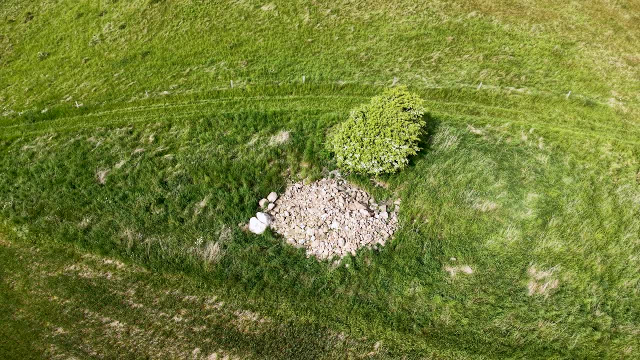 Drone footage showcases a small circular stone structure in a lush green field in Denmark. The aerial perspective highlights the stone circle surrounded by vibrant grass and rolling landscape