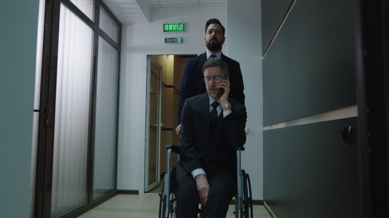 Wheelchair Manager Phoning in Office Corridor. Medium shot of disabled manager phoning while colleague pushing her wheelchair in office corridor