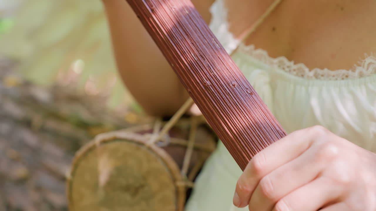 Close up of white woman with angel wing holding wooden staff under sunlight, turning it gently with talking drum beside, surrounded by soft greenery background