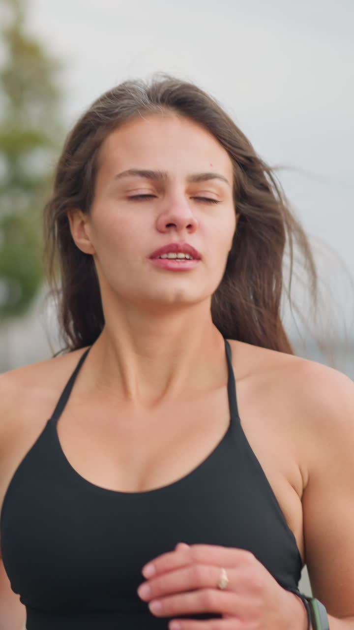 Close-up view of focused young woman in black neck top jogging with full concentration, blurred background showcasing a peaceful park setting, trees, and iron railings