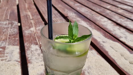 un vaso de cóctel de mojito en una cubierta de madera de arena en la playa, cerca, inclinado hacia arriba