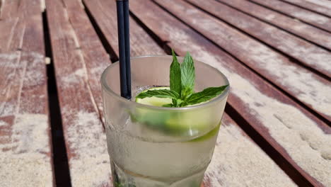 one glass of mojito cocktail on a sandy wooden deck on the beach, close up, tilt up