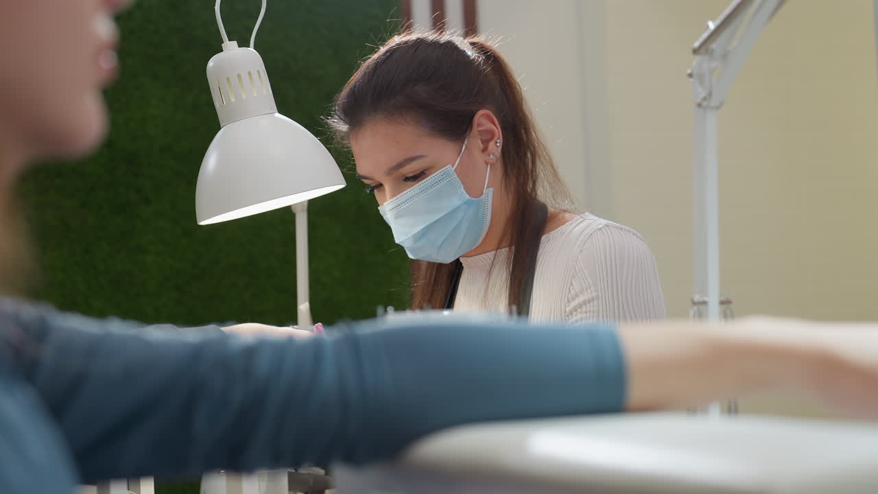 Nail technician in face mask and pink gloves concentrates while filing client nail under white lamp, as another customer rests her hand on surface in blurred background, showcasing salon detail