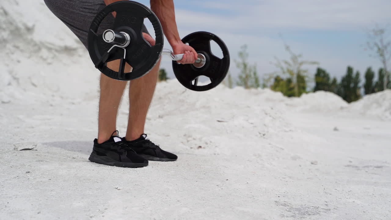 Athlete is taking heavy barbell on the natural background. Sporty man in shorts and trainers starting his workout to lift barbell outdoors. Slow motion.