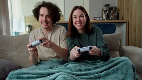 happy and confident brunette guy with curly hair plays with a joystick with his girlfriend who beats him in a video game while sitting on the couch at home