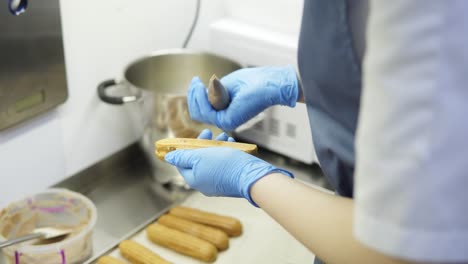 manos de confiteros irreconocibles con guantes azules llenan eclaires o profiteroles con crema batida de chocolate para terminar la preparación de deliciosos postres.