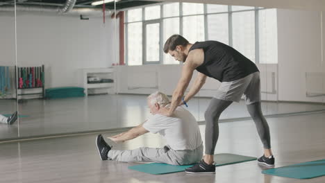 pensioner man doing stretching exercise with fitness instructor in gym together