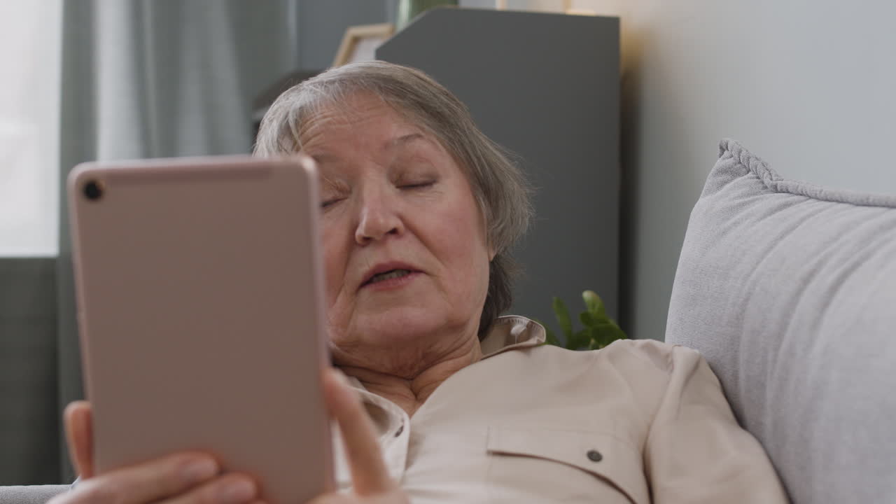 mujer mayor feliz que tiene una videollamada a través de una tableta mientras está acostada en el sofá en casa 1