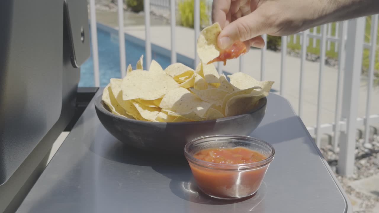 Bowl of tortilla chips and salsa at a backyard bbq, in ground pool in background. A hand picks up a chip and scoops the salsa then leaves the frame