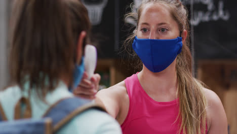 caucasian female gym receptionist in face mask taking temperature of female client