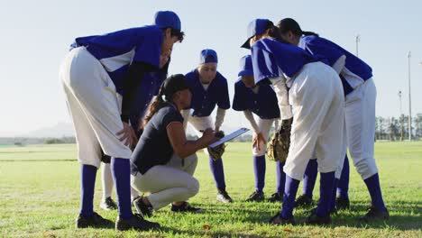 diverse group of female baseball players huddled around female coach, squatting and talking on pitch