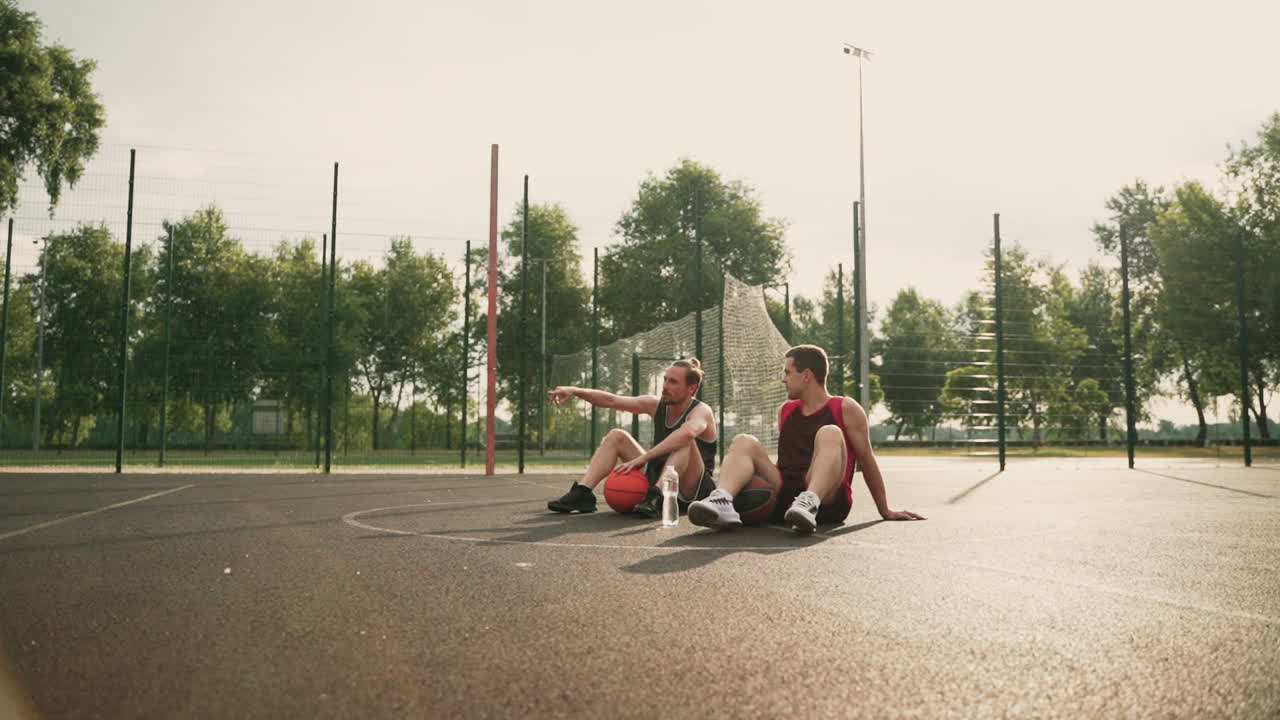 Two Happy Basketball Players Sitting In The Middle Of An Outdoor Basketball Court, Taking A Break And Talking To Each Other About Some Interesting Topic