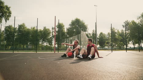 Dos-Felices-Jugadores-De-Baloncesto-Sentados-En-Medio-De-Una-Cancha-De-Baloncesto-Al-Aire-Libre,-Tomando-Un-Descanso-Y-Hablando-Entre-Ellos-Sobre-Algún-Tema-Interesante