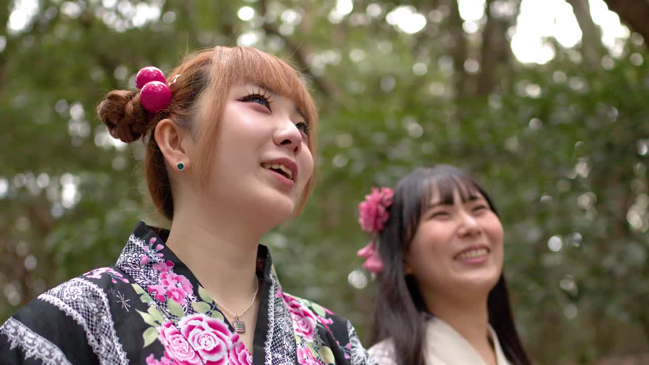 Zwei lächelnde japanische Frauen in Blumenkimonos gehen durch einen üppigen Wald in Tokio.