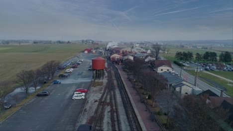 an aerial view of a train station, with a steam passenger train approaching in the distance, on a partially sunny day