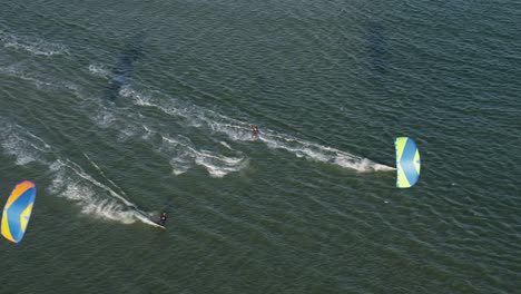 In-Dieser-Drohnenaufnahme-Von-Oben-Gleiten-Drei-Kitesurfer-über-Das-Meerwasser,-Während-Der-In-Der-Mitte-Akrobatische-Tricks-Vorführt