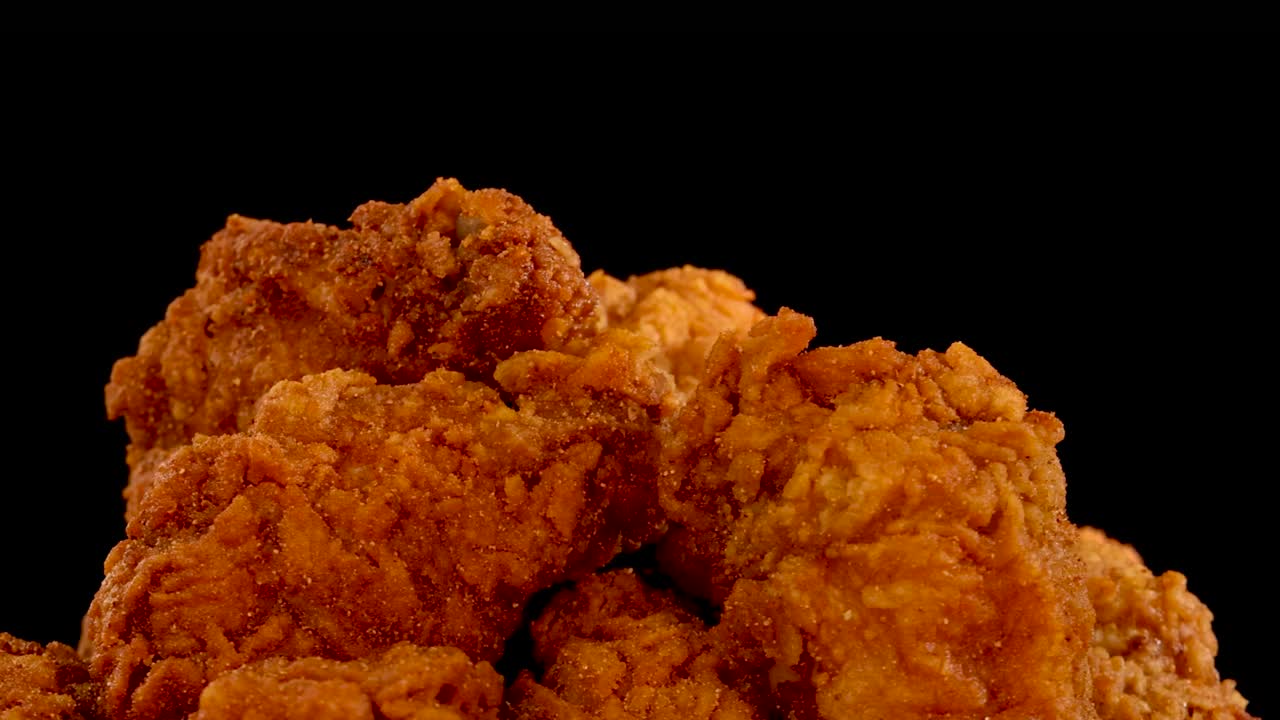 Crispy, golden fried chicken pieces are shown in close-up against a black background, with slow motion zoom revealing texture and seasoning in dramatic lighting