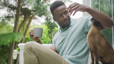 Happy-african-american-man-sitting-with-cat-and-drinking-coffee-in-garden