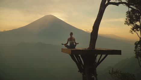 mujer rubia en pose de yoga en la plataforma de la cabaña del árbol, vista impresionante del monte agung, puesta de sol