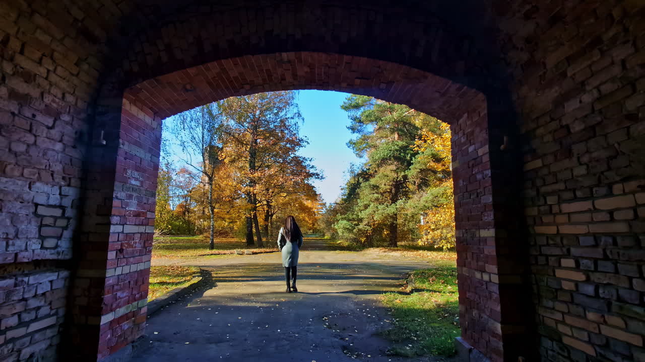 Person walks through an old brick tunnel into a sunlit autumn park. Lielvarde, Latvia