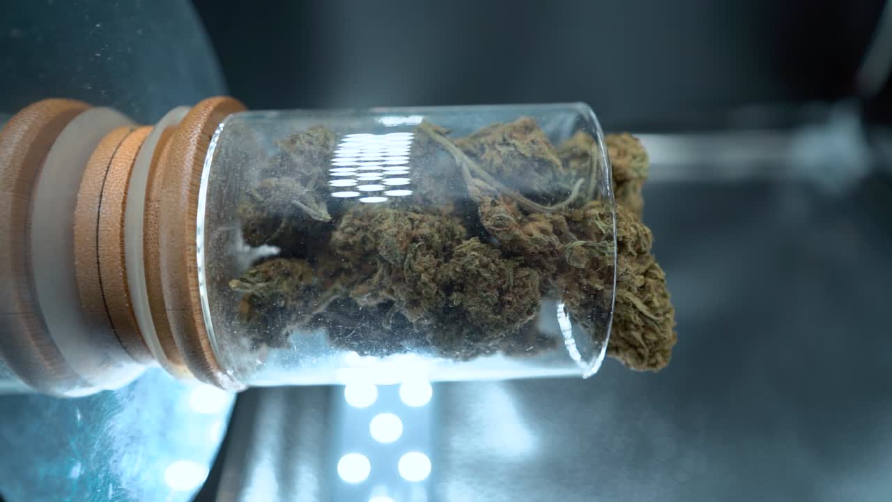 Grønne tørrede cannabisblomsterstande i et vertikalt nærbillede. En bunke tørrede cannabisplanter i et skinnende glas, placeret på et reflekterende roterende stativ, med studiebelysning, i en filmisk slowmotion 120 fps video.