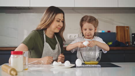 mother-looks-at-daughter-in-apron-cracking-egg-into-bowl