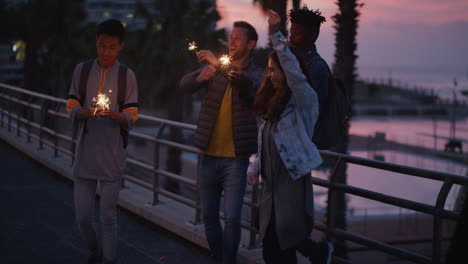 Porträt-Junger-Multiethnischer-Freunde,-Die-Winkende-Wunderkerzen-Tanzen-Und-Die-Silvesterfeier-Am-Abend-Der-Freundschaft-In-Der-Stadt-Am-Meer-Genießen