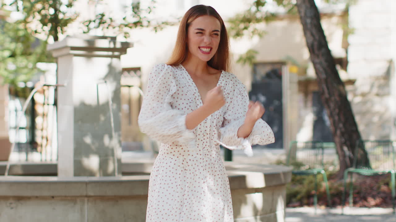 색 폴카 도트 드레스를 입은 한 젊은 여성이 웃으며 마을 한가운데의 분수 앞에서 어깨를 뒤집어보고 있습니다.